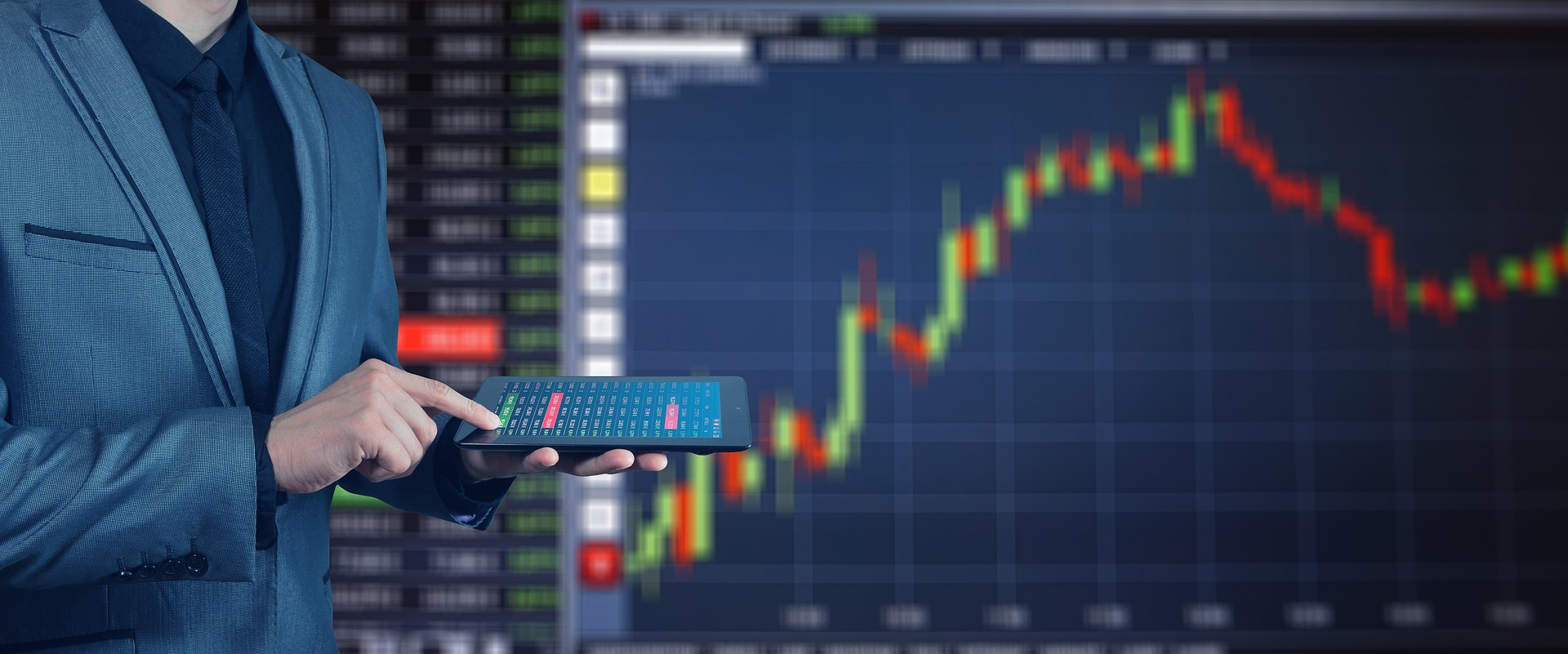 Person stands with a tablet in front of a large screen showing the stock market performance