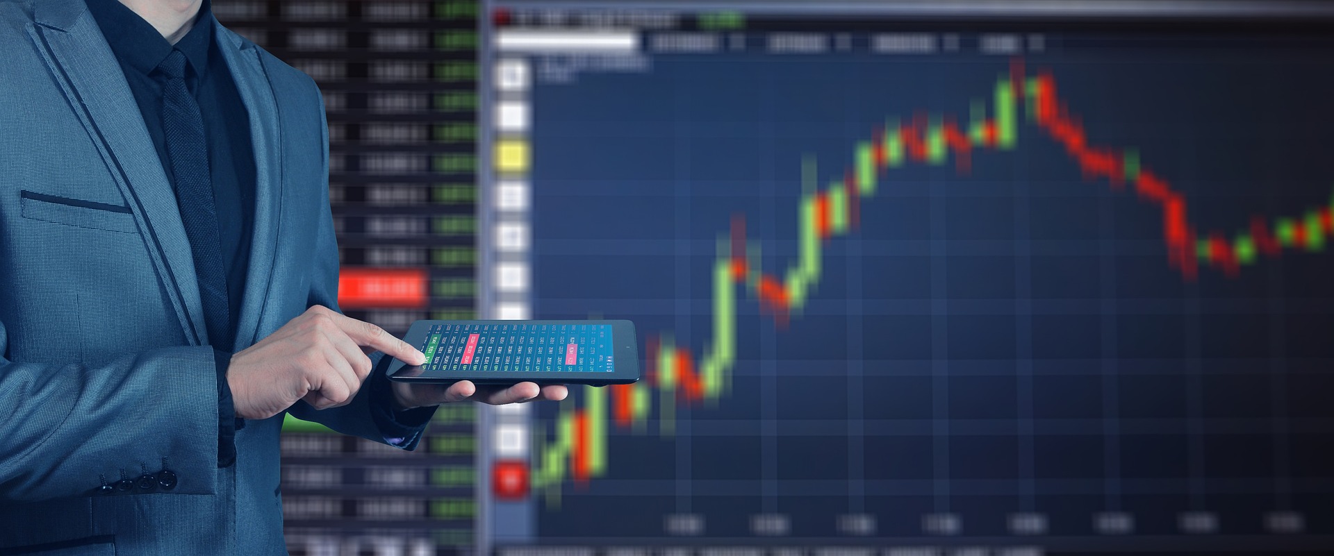 Person steht mit Tablet vor einem grossen Screen mit Börsenverlauf